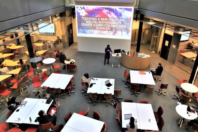 Etualalla kestävyyskurssin osallistujia istumassa pöytien ääressä Helsingin yliopistolla. Taustalla luennoitsija ja dia, jossa lukee "Creating a new discipline specific sustainability course (computer and data sciences)".