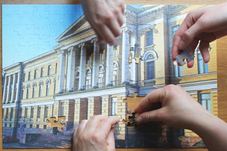 Käsiä kokoamassa yliopiston päärakennusta esittävää palapeliä.