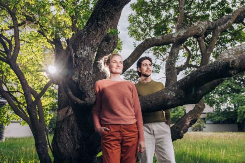 TWO PEOPLE ARE STANDING UNDER A TREE, LOOKING HOPEFULLY AHEAD.
