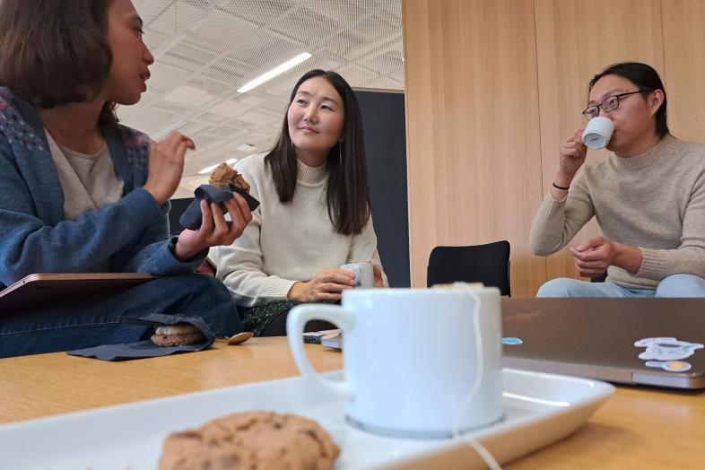 Three students sitting at a table drinking tea/coffee and eating biscuits.