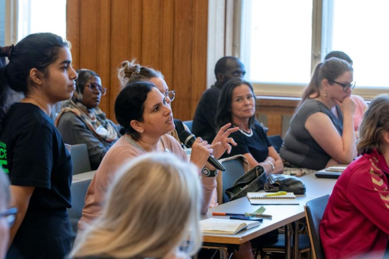 A group of people sitting in a lecture hall. In the middle a person, who is talking into a microphone.