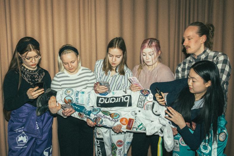 Viisi puhelinta selailevaa opiskelijaa pitää sylissä kuudetta, jolla myös puhelin kädessä.